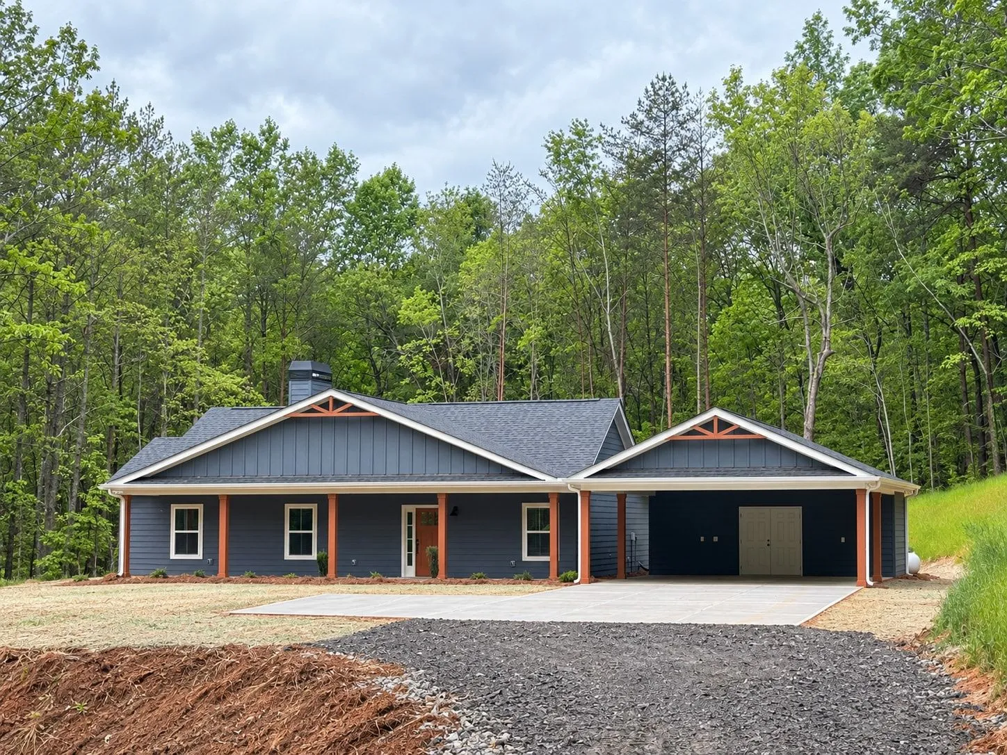 Single-story blue house with a long covered front porch on wood posts and an attached carport, set against the woods